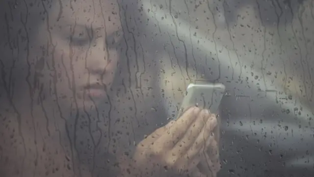 فتاة تتصفح هاتفها المحمول خلف نافذة زجاجية