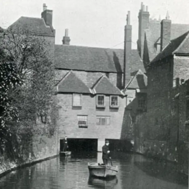 Un edificio medieval que cruza un río, en blanco y negro. Se ven dos apostadores en el agua, con árboles en la muralla que la bordea.