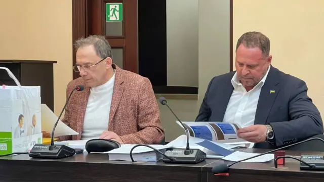 У залі засідань за столом із мікрофонами сидять Єрмак і Фомін і читають документи. На столі — папери та папки, позаду видно двері з табличкою евакуаційного виходу.