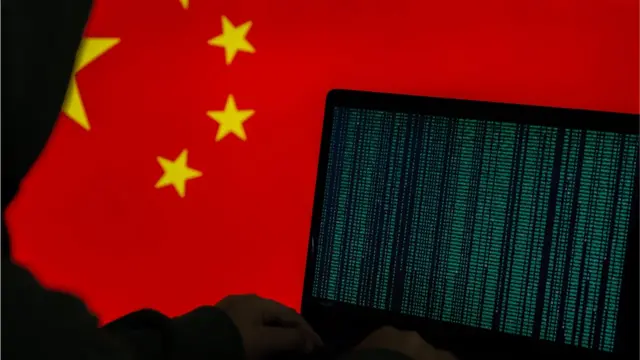 Stock image of hooded person working on computer in front of a Chinese flag