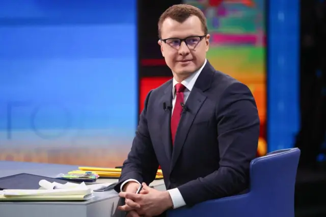 Павел Зарубин в костюме сидит вполоборота в кресле, слегка улыбаясь