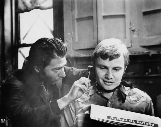 Ratso Rizzo (Dustin Hoffman) le corta el pelo a Joe Buck (Jon Voight), mientras Buck escanea una columna personal.