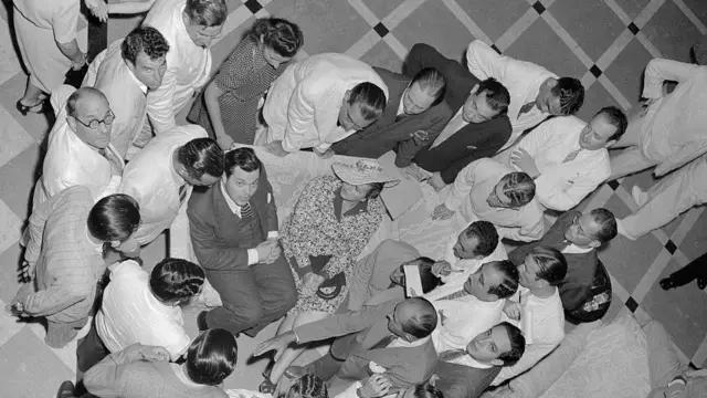 Welles sentado e olhando para cima, rodeado por repórteres em pé