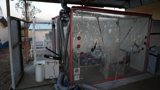 Un lit se trouve à l'intérieur d'une cabine en plastique. Les tuyaux sortent de la cabine.