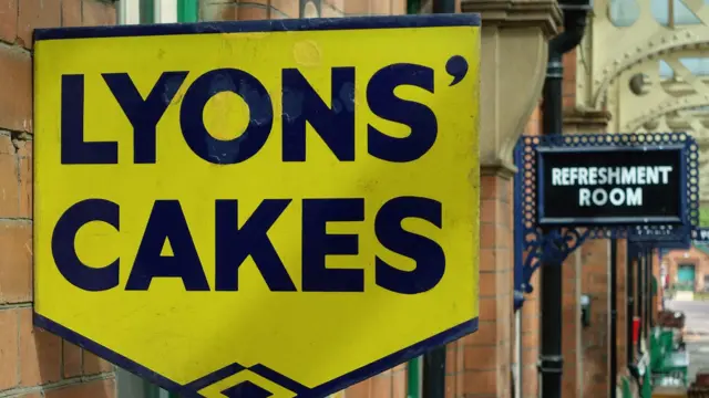 Улица, желтая табличка с названием компании «Lyons' Cakes» на первом плане. Название написано черными заглавными буквами. На заднем плане название другой компании и вид улцы.