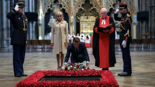 キリスト教の寺院の床にはめ込まれ、周囲を赤い花に囲まれた「無名戦士の墓」の石板がある。マクロン氏がその前にしゃかみ、花輪を供えている。そばにはブリジット氏と司祭、敬礼した警備員2人が立っている。背後には聖歌隊の姿も見える