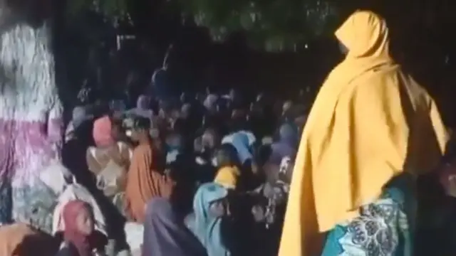 A screengrab from a video showing a crowd of mainly and children, mostly from behind, as dem sit under a tree afta dem gain freedom from Boko Haram captivity. One of di women dey stand to di right of di image and wear a yellow headscarf.