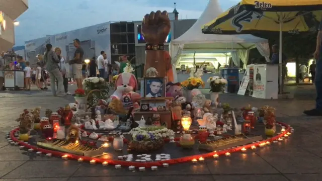 Дрвена песница усмерена ка небу испред које стоји фотографија Давида Драгичевића окружена десетинама упаљених свећа. Август 2018, Бањалука.