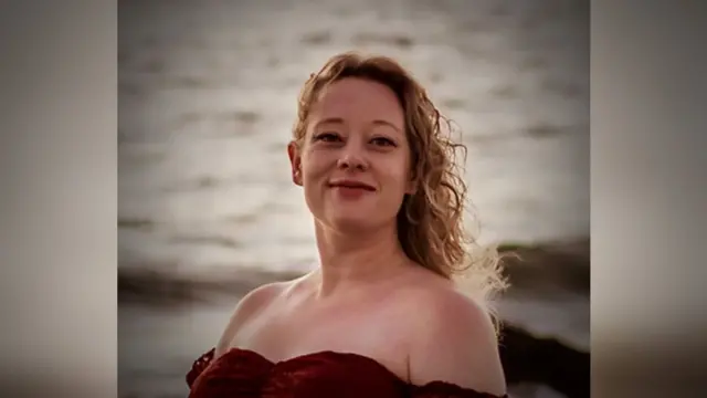 Renee Good tem cabelos longos, loiros e cacheados e veste uma blusa vermelha de ombro a ombro. O oceano está ao fundo.