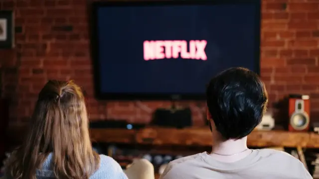 De costas, duas pessoas, umacomo fazer aposta lotofácil onlinecabelo loiro e comprido e outracomo fazer aposta lotofácil onlinecabelo preto curto, assistem sentadas a uma TV que exibe a palavra Netflix