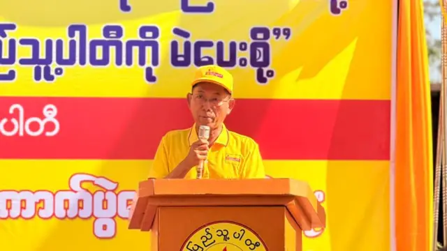 အဝါရောင်ဆိုင်းဘုတ်နောက်ခံနဲ့ စင်မြင့်ပေါ်မှာ ပါတီဦးထုပ်အဝါ၊ တီရှပ်အဝါဝတ်ပြီး မိန့်ခွန်းပြောနေတဲ့ ပြည်သူ့ပါတီဥက္ကဋ္ဌ ဦးကိုကိုကြီး 