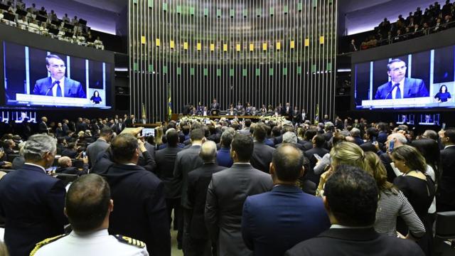 Bolsonaro aparece em duas telas, discursando a parlamentares no plenário da Câmara