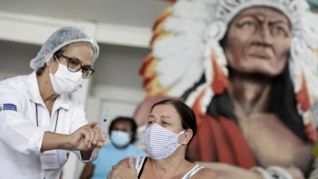 Profissional vacina mulher sentada; atrás, é possível ver grande escultura carnavalescabaixar lampions betsum cacique