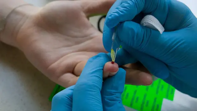 Un sanitario con guantes azules recoge una muestra de sangre del dedo de un paciente.