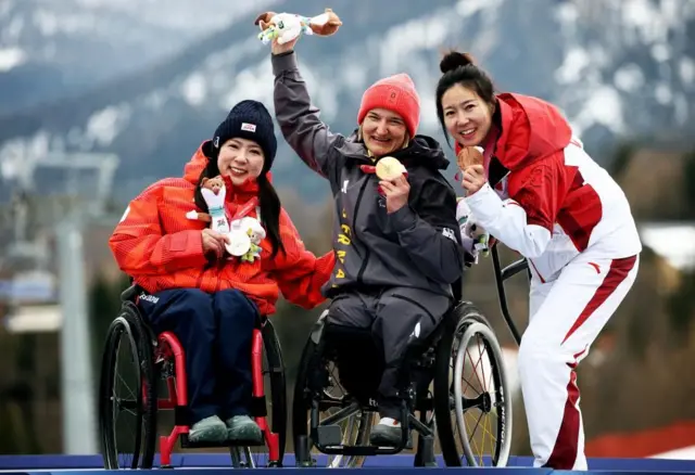 表彰台の上で3人が横に並んでいる。全員メダルを胸の前に持ち、笑顔を見せている。村岡とフォルスターは車いすに座り、劉は杖を使って立っている