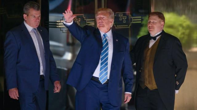 Trump in blue suit with blue tie in front of glass doors, between man in blue suit and door man in black suit with gold vest.