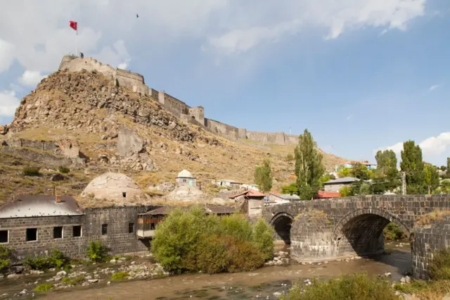 Kars Kalesi, üstüne Türk bayrağı, etrafta çeşitli eserlerden kalıntılar ve ağaçlar