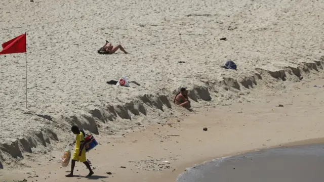 Ambulante caminha na areafutebol de 5praia no Riofutebol de 5Janeiro, com duas mulheres pegando sol ao fundo