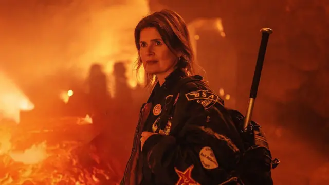 Sophie Aldred in Doctor Who in 2022, dressed in her jacket, stood inside a film set made to look like a volcano, near a stream of lava.