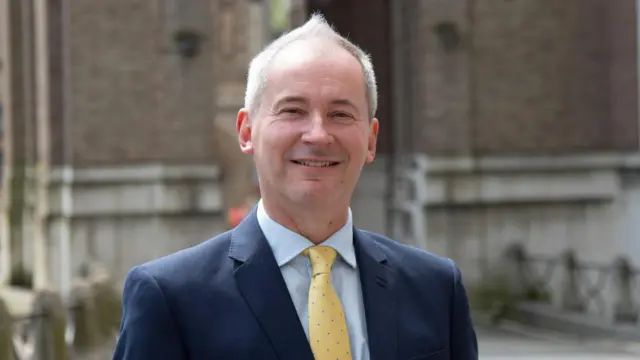 El concejal Stephen Williams se encuentra frente al Ayuntamiento de Bristol con traje azul y corbata amarilla. Tiene el pelo corto y canoso y sonríe a la cámara.