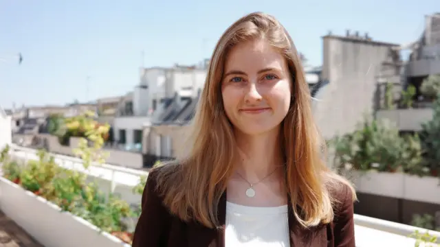 Emma Fourreau egy napfényes erkélyen áll, növényekkel a háttérben. Barna dzsekit visel, amely nyitva van, alatta fehér felső és egy nyaklánc látható. Vállig érő szőke haja van, és mosolyog.