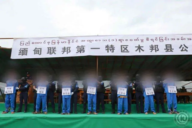 လည်ပင်းမှာ တရုတ်စာရေးသားထားတဲ့ ကတ္ထူချိတ်ဆွဲပြီး အပြာဝမ်းဆက် အကျဥ်းသားဝတ်စုံနဲ့ အကျဥ်းသားတွေ၊အမဲဝမ်းဆက် ကိုးကန့်ရဲဝတ်စုံနဲ့ ကိုးကန့်ရဲတပ်ဖွဲ့ဝင်တွေ စင်တစ်ခု အပေါ်မှာ ရပ်နေပုံ