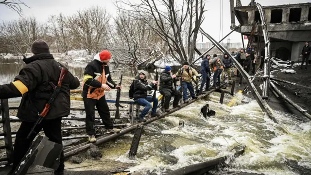 यूक्रेन सिविल डिफेंस के सदस्य 1 मार्च 2022 को कीएव में एक उड़ा दिए पुल के पास हथियार एक दूसरे को बढ़ाते हुए