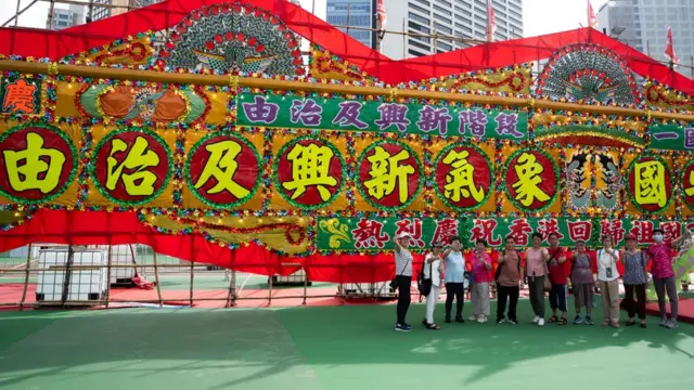 香港維多利亞公園足球場上一群長者在親建制團體慶祝「七一」活動的「由治及興」大型花牌前拍照留念(30/6/2023)