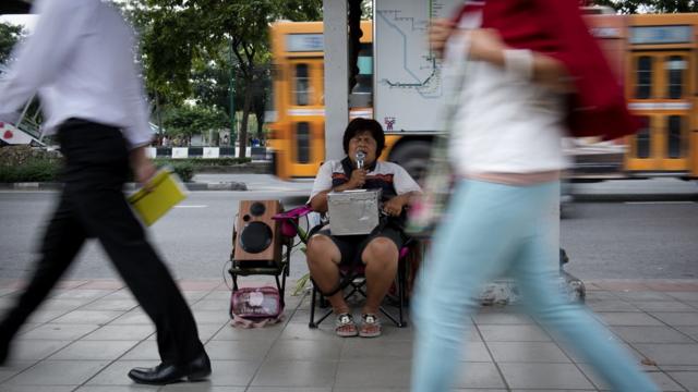 ภาพคนตาบอดหญิงจับไมค์นั่งร้องเพลงบนทางเท้า โดยมีหีบใส่เงินวางอยู่บนตัก