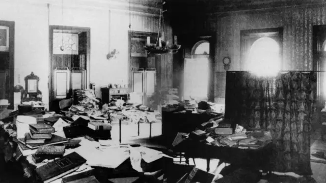 Foto antiga mostra livros e papéis espalhados por cômodo