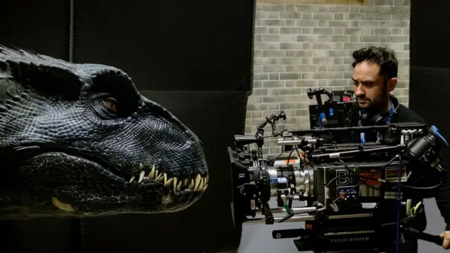 Um cinegrafista segura uma camera apontada para um robôaposta online loteria dos sonhosdinossauro durante a filmagem do longa 'Jurassic World: Reino Ameaçado'