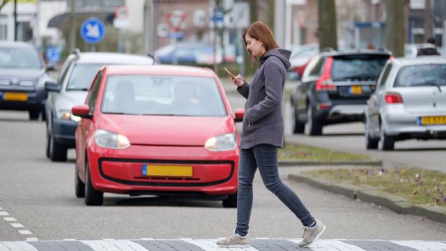 Una mujer mira su celular mientras cruza la calle y no ve los autos.