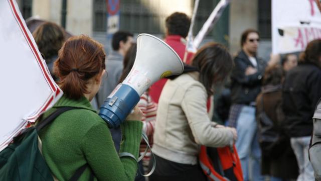 Diversos estudantes aparecem em rua com cartazes, uma delas segurando um megafone