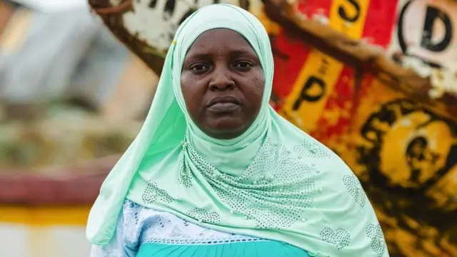 Adjointe au maire portant un foulard vert vif devant un bateau en bois.