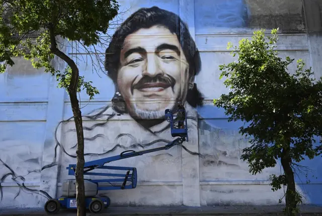An artist uses a crane to paint a large mural of Maradona on the side of a building