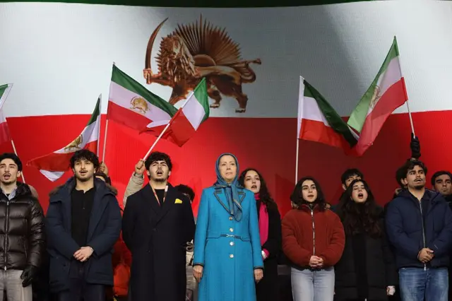 Recavi yanında İran bayrakları sallayan gençler ile sahnede. Üzerinde mavi bir takım var, başı ise kapalı.