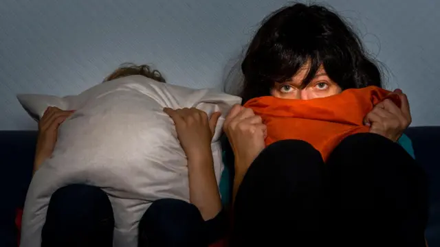 Duas mulheres vendo televisão sentadas na cama. Uma delas se cobre totalmente com uma almofada e a outra leva o cobertor à boca, mas continua assistindo atentamente com os olhos assustados