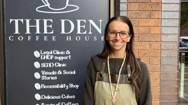 Claire Naylor lleva un jersey negro, un delantal y gafas, de pie frente a un gran cartel de The Den Coffee House, que enumera sus servicios. Estos incluyen una clínica Send, una tienda de accesibilidad y apoyo con planes de educación, salud y atención.
