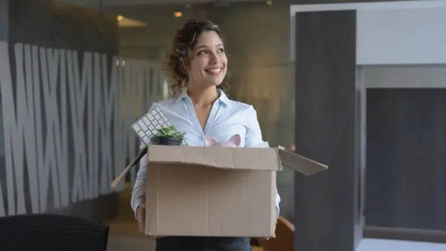 Una mujer sonríe mientras sostiene una caja llena de cosa de oficina.