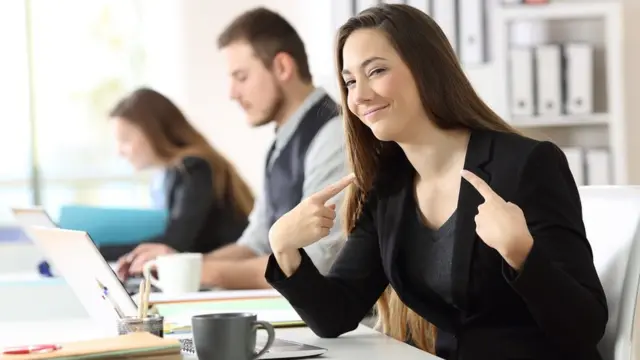 কর্মক্ষেত্রে ডেস্কে বসা খুবই গর্বিত এক ব্যবসায়ী নারী নিজের দিকে দুই আঙুল তুলে বুঝিয়ে দিচ্ছেন তিনিই সর্বশ্রেষ্ঠ।