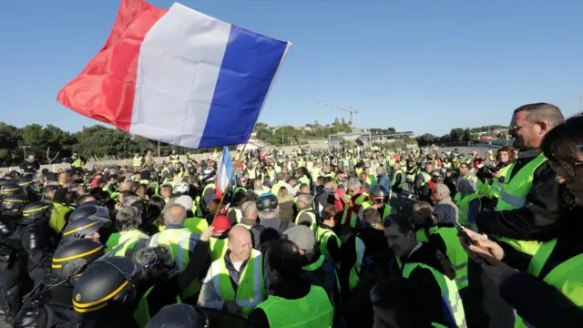 No Sul da França, polícia francesa tenta conter manifestantes vestidosceara x coritiba palpitecoletes amarelos; um deles hasteia uma bandeira da França