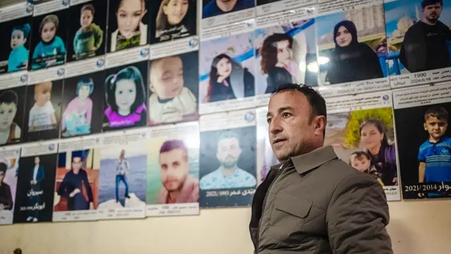 Внутреннее помещение со стеной, плотно завешанной рядами отпечатанных портретных фотографий; каждая фотография закреплена на карточке, под которой расположены строки текста, часть которых написана на арабском. На фотографиях запечатлены взрослые и дети. На переднем плане стоит Бакр Али, слегка повернувшись вбок, в утепленной куртке на молнии. 