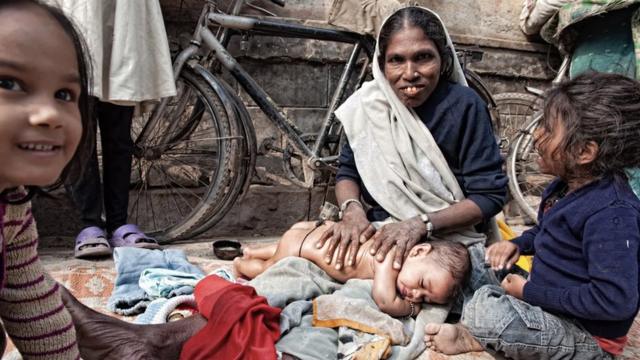 Una mujer masajea a un bebé con los ojos cerrados en una calle, con dos niños mayores sentados a su lado