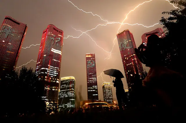 A bolt of lightning crosses the sky as people look at buildings displaying a light show on the eve of the 100th anniversary of the Chinese Communist Party in Beijing