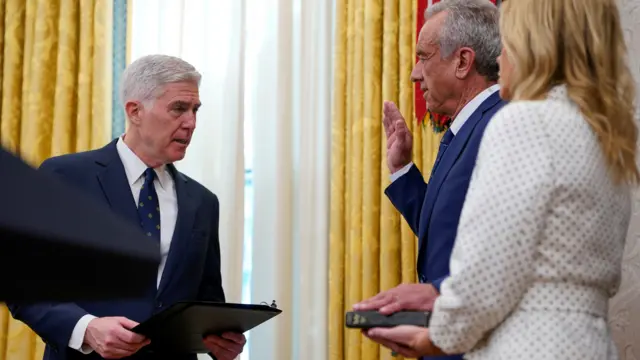 Кеннеди приносит присягу, одна рука в согнутом положении поднята вверх, другая лежит на Библии, которую держит женщина в белой одежде. Это его жена Шерил Хайнс. Рядом стоит мужчина с папкой в деловом костюме: он диктовал текст присяги.