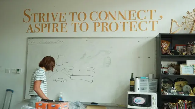 一名短髮女子在白板前，牆上以大字寫着「STRIVE TO CONNECT, ASPIRE TO PROTECT」，周圍擺放各種獎盃與紀念品。