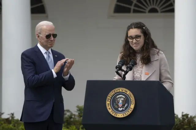 US President Joe Biden applauds as Mia Tretta, a victim of gun violence, speaks during an event in the Rose Garden of the White House in April 2022. BIden is wearing a navy suit and blue tie and Mia is wearing a pink dress and coat and stnding in from of a podium