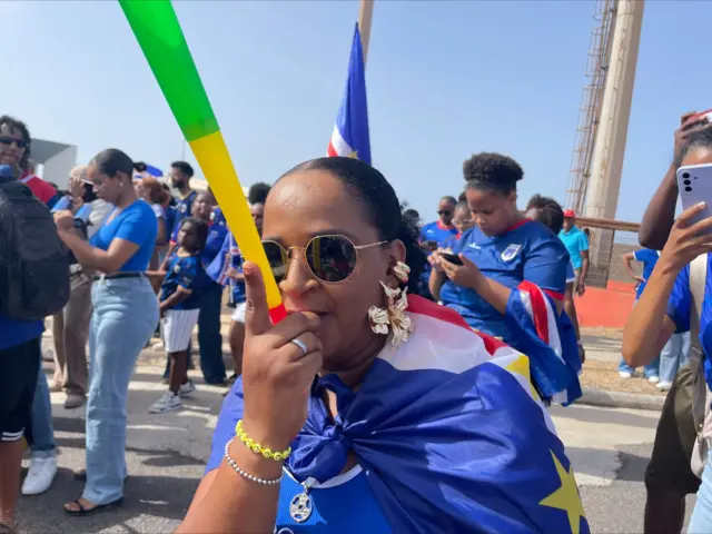 Une femme soufflant dans un vouvouzela, avec en arrière-plan plusieurs supporters.