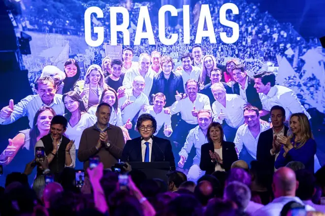 Milei e apoiadores posam para foto com painel ao fundo escrito Gracias