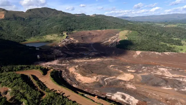 Visão da barragem da mina CórregoFeijão após o rompimento; a estrada onde estava o caminhãoAna Paula fica à direita do entroncamento"v", tomada parcialmente pela lama